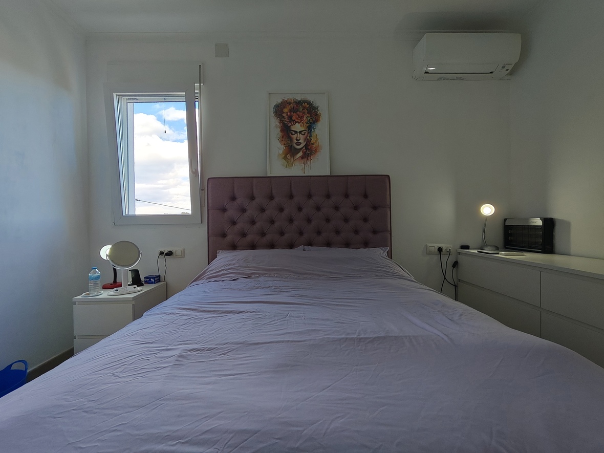 Bedroom featuring a mauve tufted headboard, white minimalist furniture, and an air conditioning unit. A portrait hangs above the bed near a window with natural light.