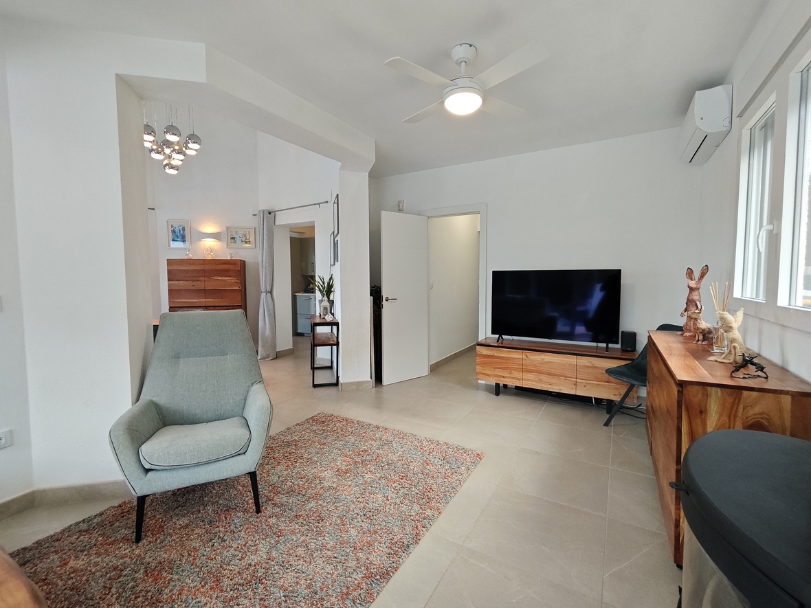 Bright living area featuring a modern armchair, natural wood media console, and flat-screen TV. Equipped with a ceiling fan, air conditioning, and neutral tiled flooring.