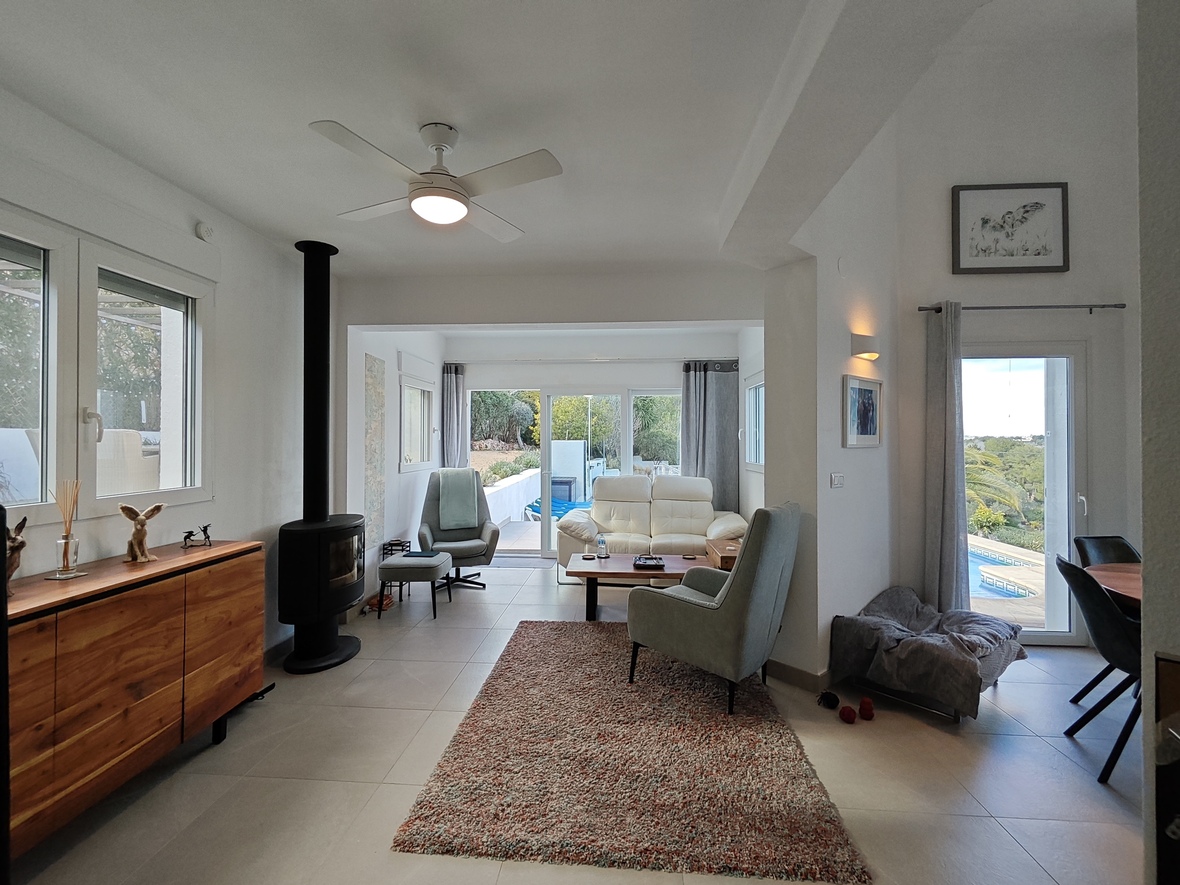Bright living room featuring a black wood-burning stove, wooden sideboard, and ceiling fan. Large glass doors offer direct access to the terrace and pool area.