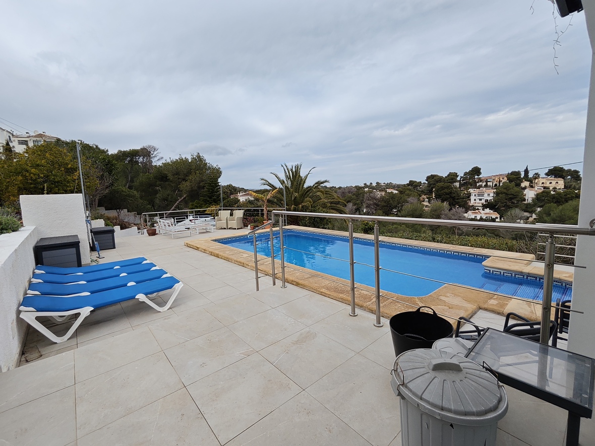 Elevated terrace featuring a rectangular swimming pool with stone borders, blue sun loungers, and stainless steel railings overlooking a wooded residential valley.