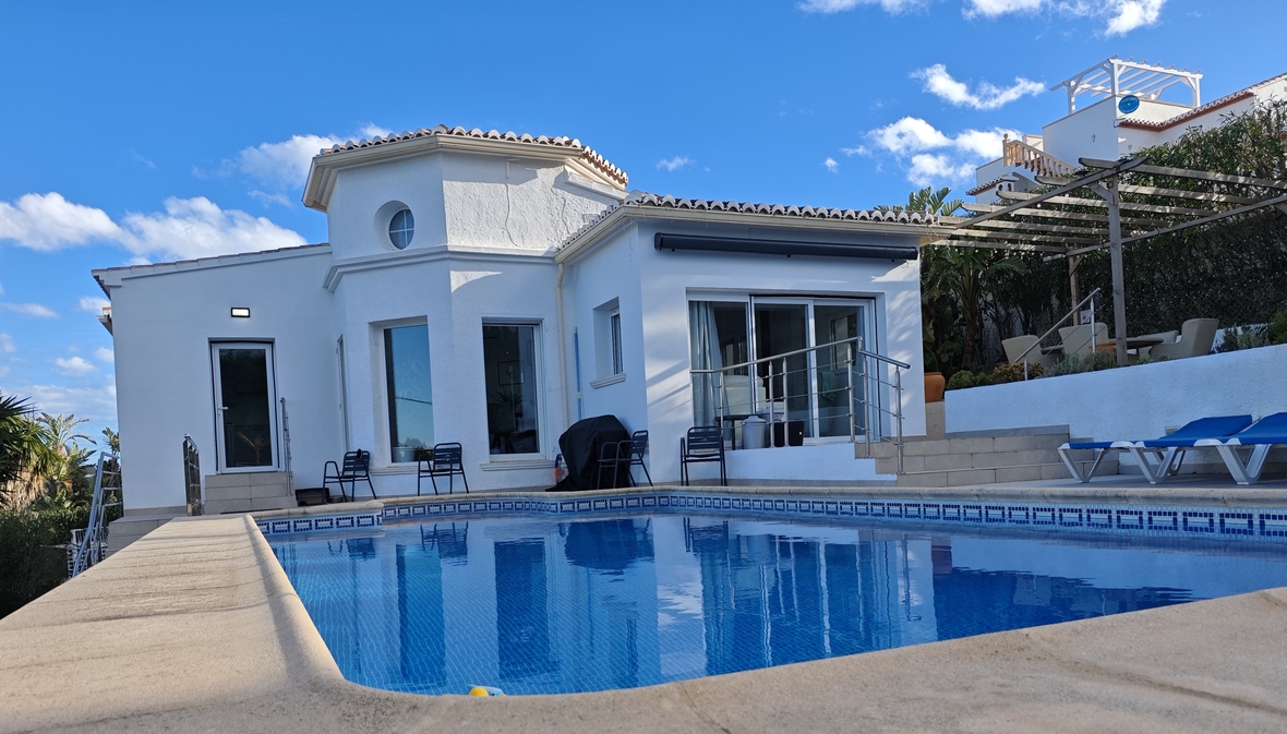 Exterior view of a white Mediterranean villa featuring a private swimming pool, sun terrace with blue loungers, and a wooden pergola.