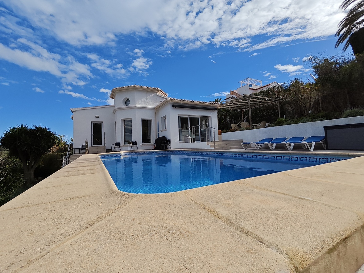 Exterior view of a white villa featuring a private swimming pool with blue tiling, stone terrace, and sun loungers under a clear sky.