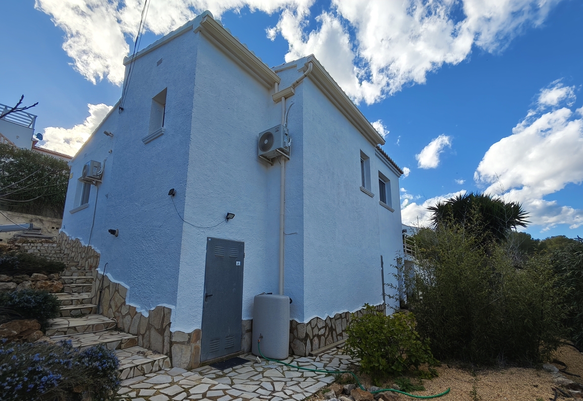 Exterior view of the villa featuring white textured walls, stone-clad base, and external utility units under a bright blue sky with scattered clouds.