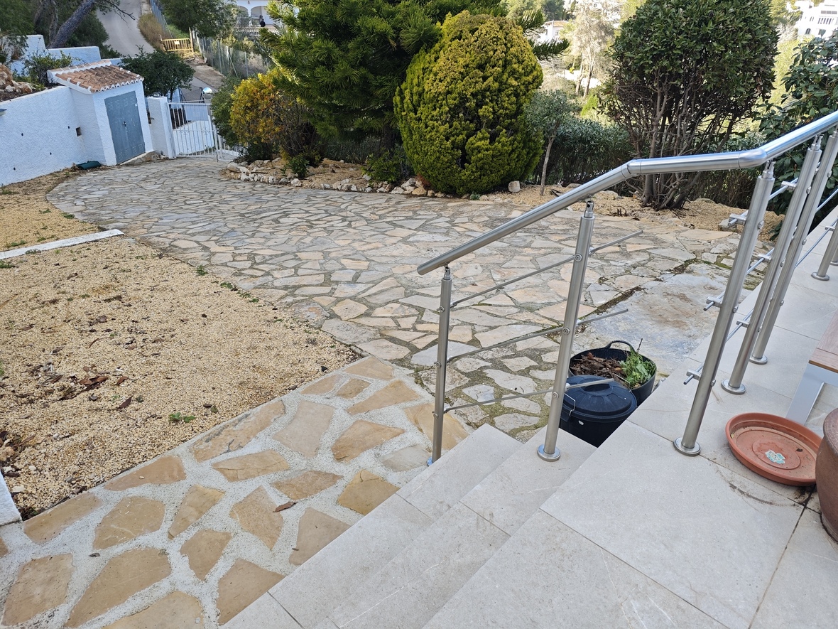 Exterior view showing a stone-paved driveway surrounded by Mediterranean vegetation and a staircase with a stainless steel railing.