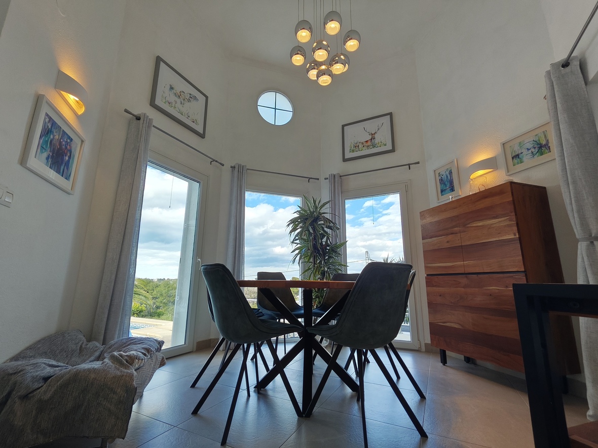 High-ceilinged dining area featuring a modern chrome sphere pendant, a circular clerestory window, and a wooden sideboard. Large glass doors provide ample natural light.