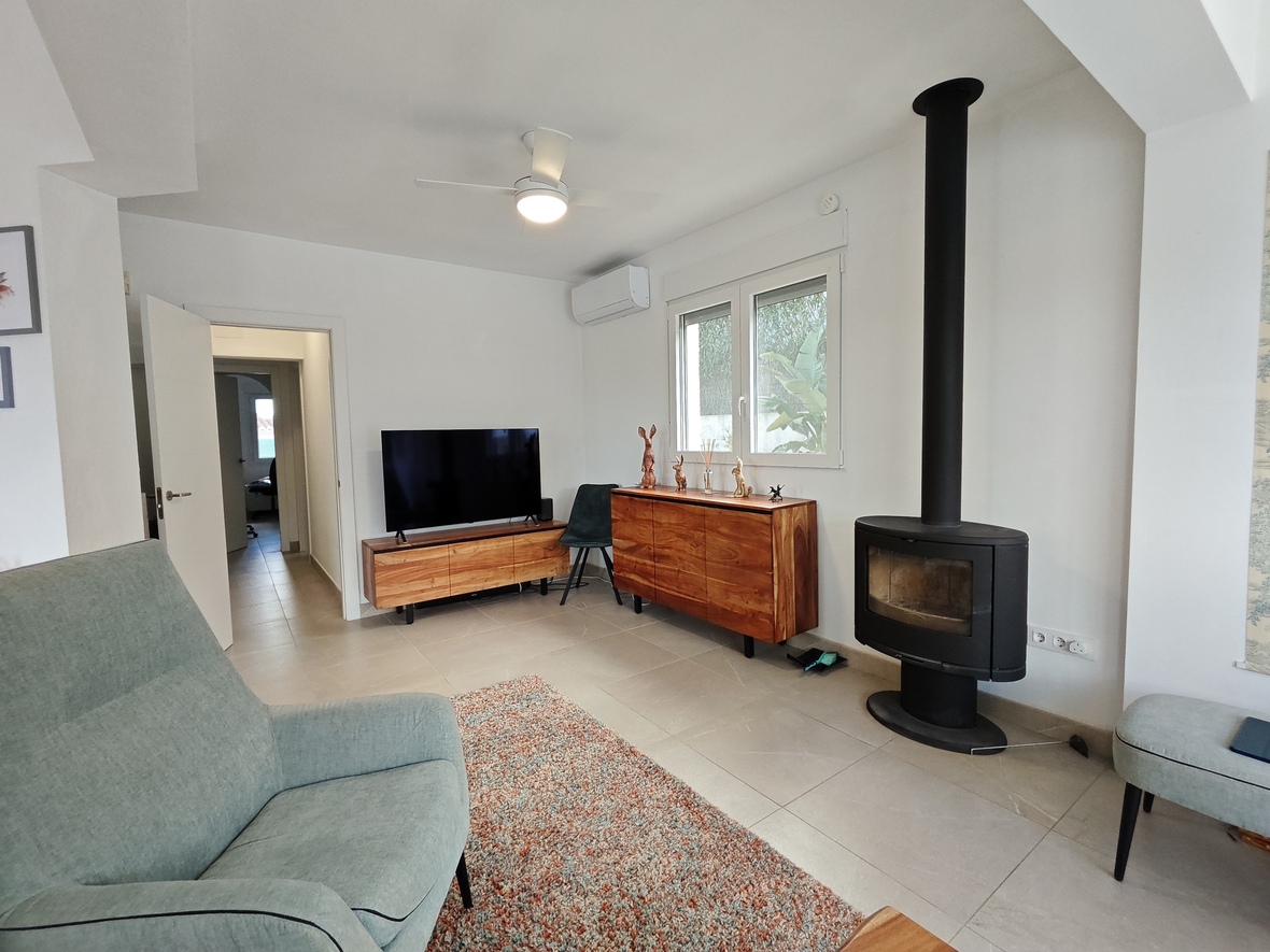 Living area featuring a modern black wood-burning stove, solid wood sideboards, and tile flooring. Natural light enters through a window overlooking garden foliage.
