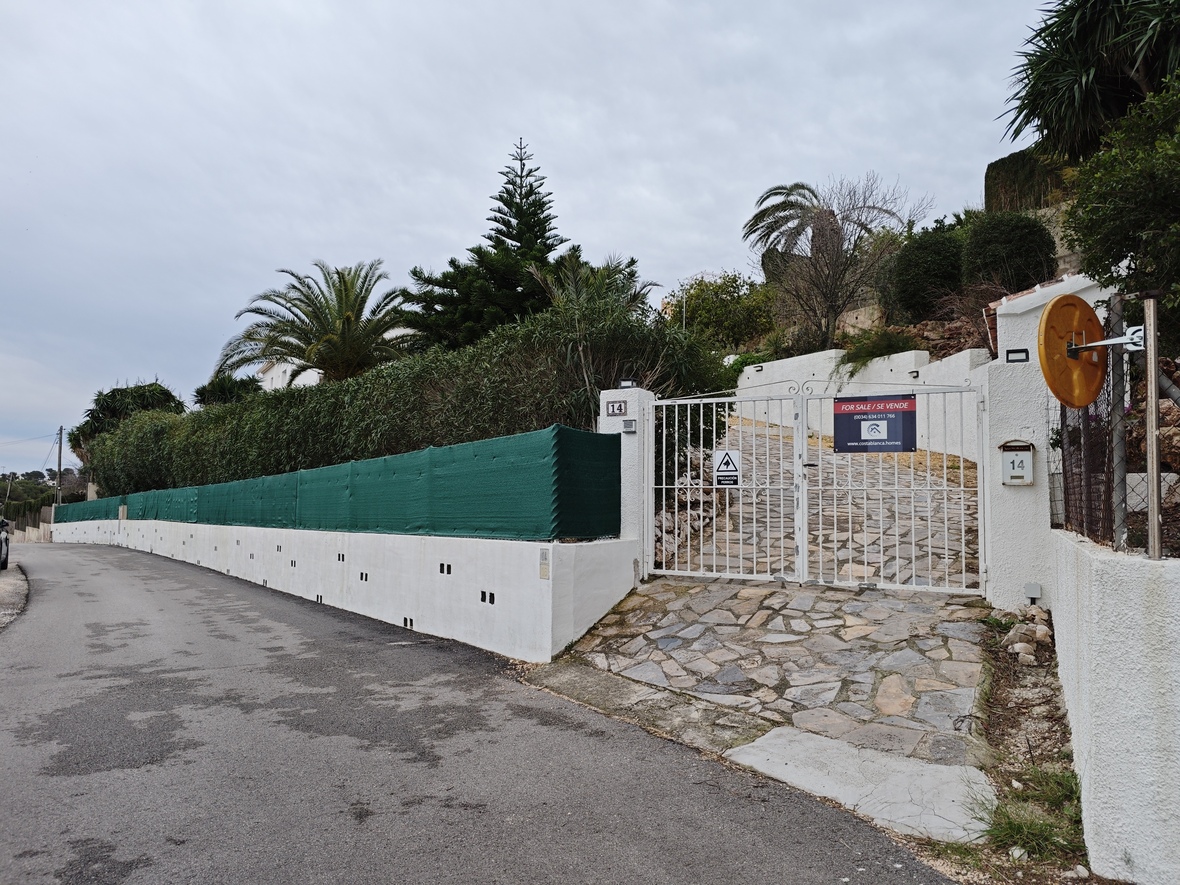 Main entrance featuring a white metal gate, a natural stone-paved driveway, and a perimeter wall with privacy screening and mature trees.