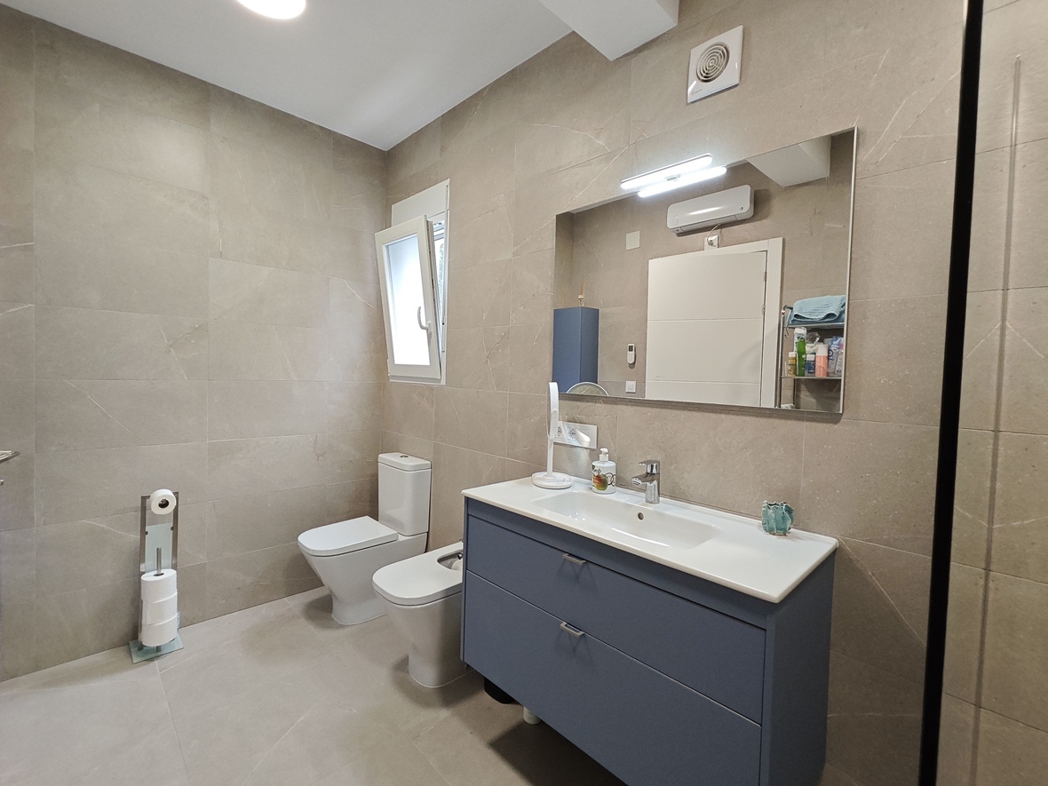 Modern bathroom featuring beige stone-effect tiling, a blue floating vanity with integrated sink, and a matching toilet and bidet set. Includes a large mirror with LED lighting.