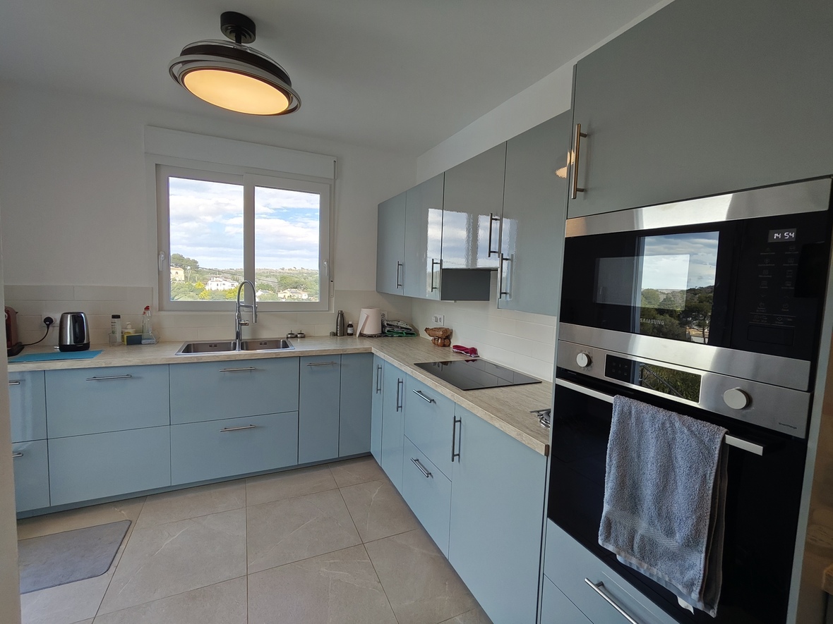 Modern kitchen featuring light blue cabinetry, wood-effect countertops, and integrated Samsung appliances. A large window provides natural light and landscape views.
