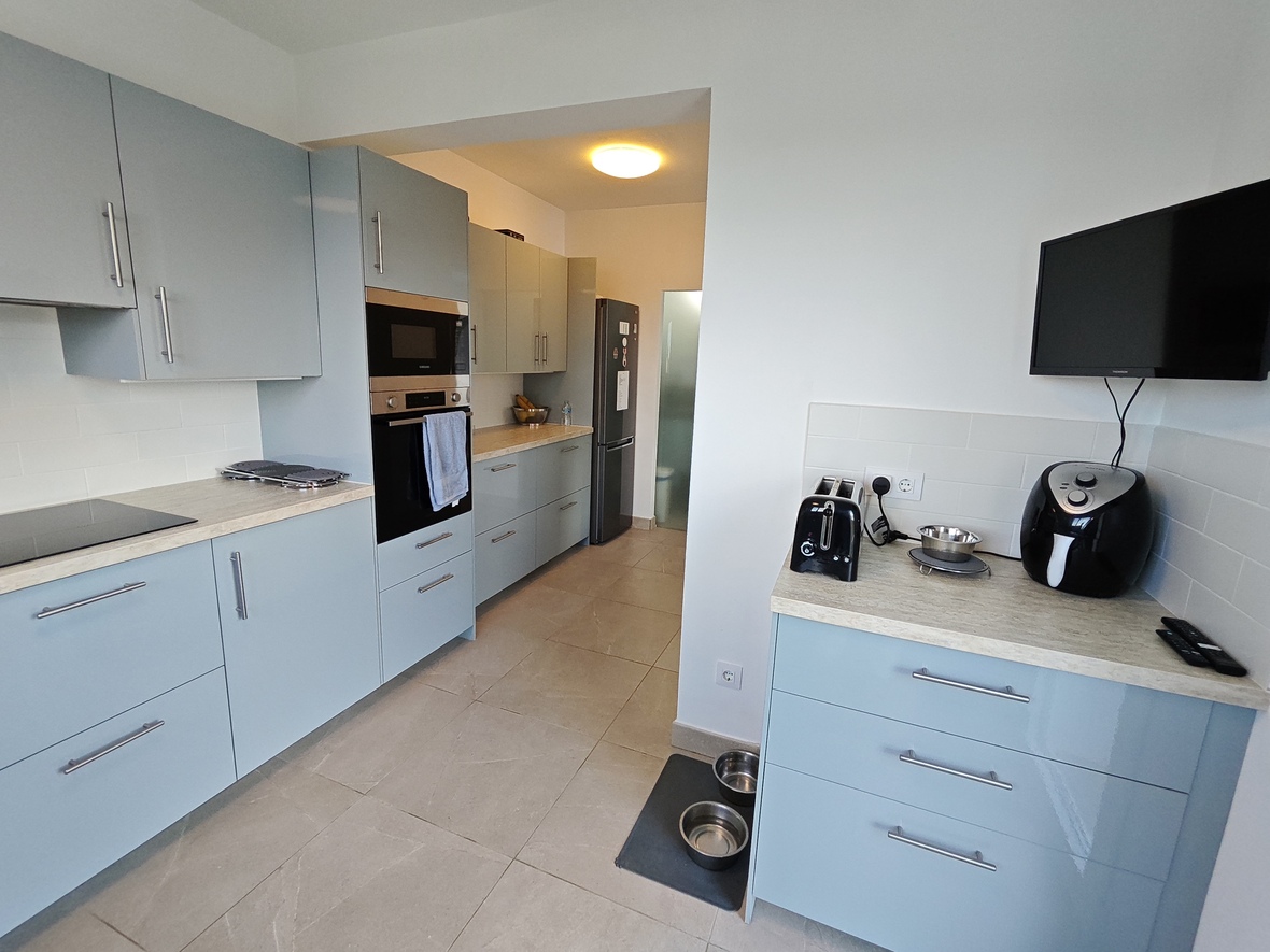 Modern kitchen with light blue cabinetry, integrated oven and microwave, stone-effect countertops, and a wall-mounted television. Grey floor tiling completes the space.