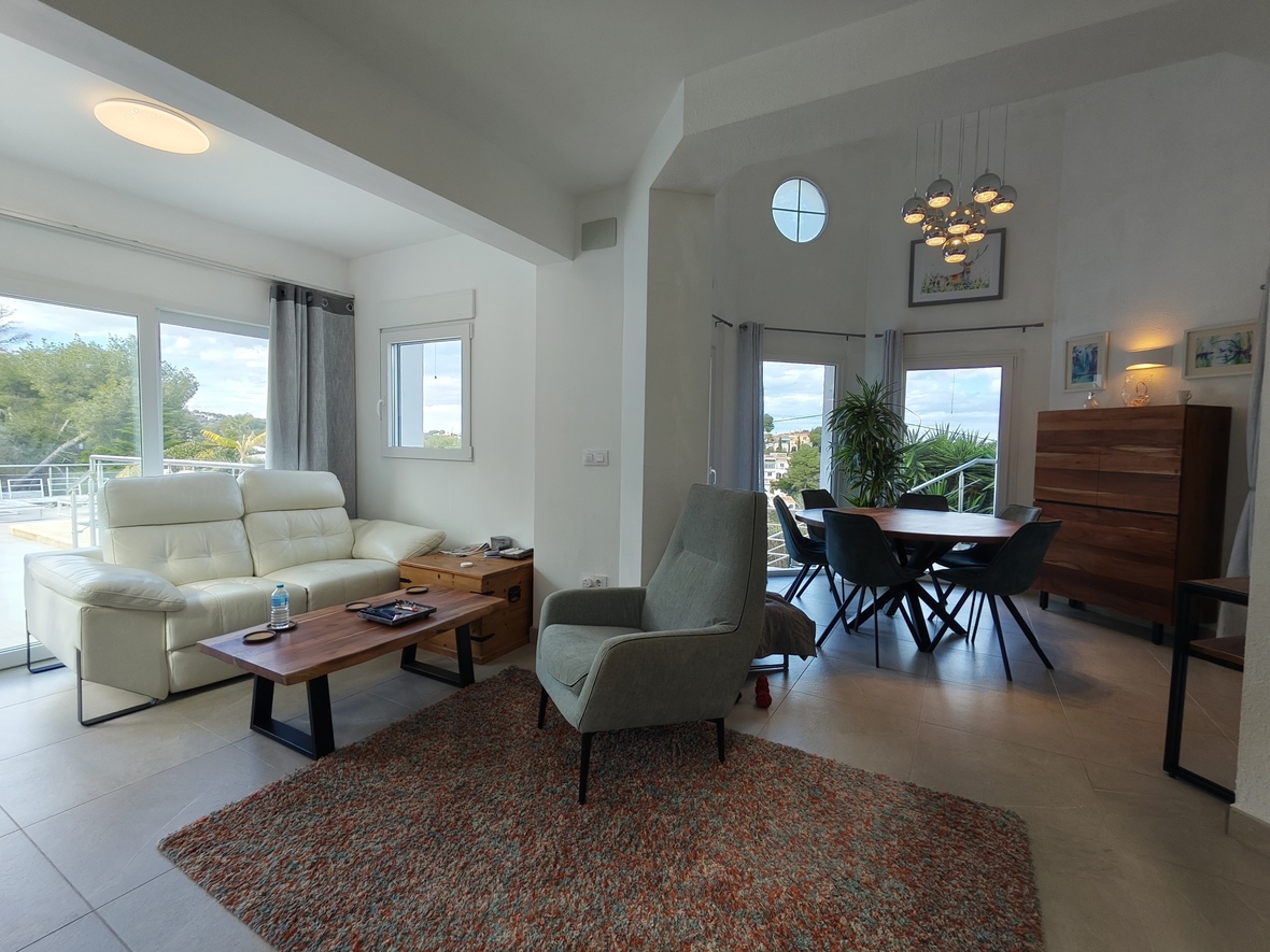 Open-plan living and dining space with white leather seating, wooden accents, and a modern cluster pendant. Large glass doors lead to a terrace with landscape views.