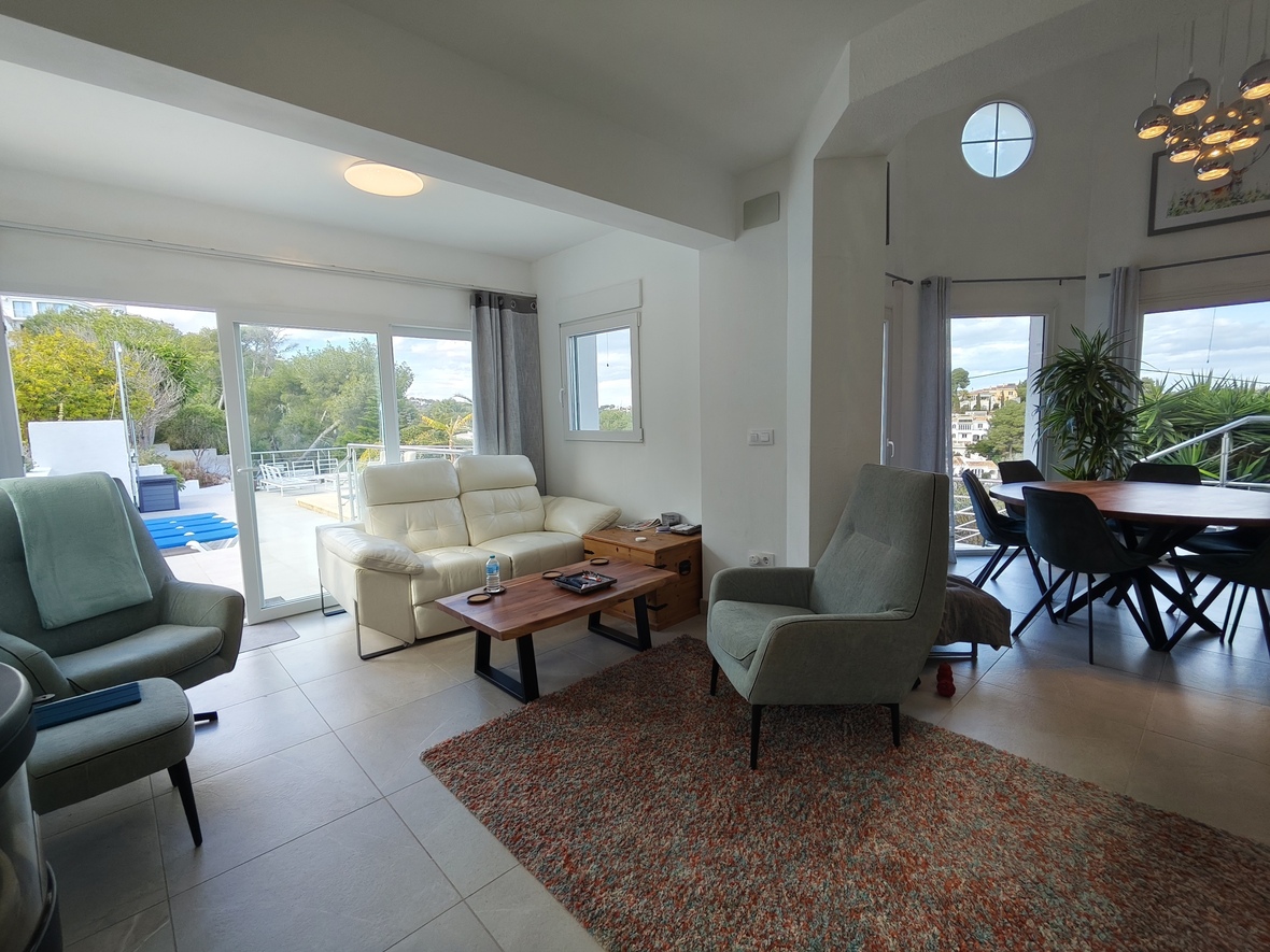 Open-plan living area featuring a white leather sofa, high ceilings with a porthole window, and large sliding doors leading to a terrace and pool.