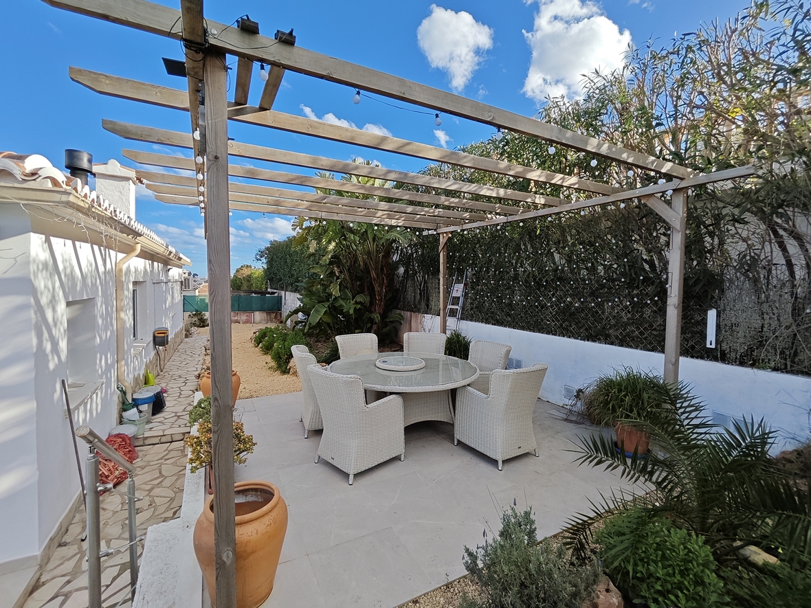 Outdoor dining area featuring a wooden pergola with string lights, a round glass-top table, and white wicker chairs set on a tiled patio.