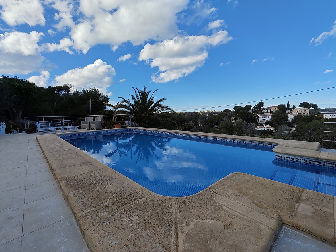 Outdoor swimming pool with blue mosaic tiling and stone borders, overlooking a Mediterranean landscape with palm trees and residential views.