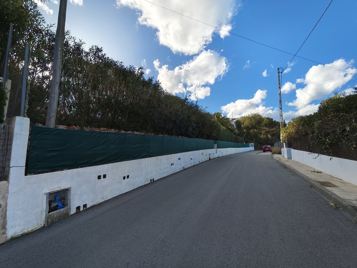 View of the residential access road featuring a white perimeter wall with green privacy screening and mature hedges under a bright, cloudy sky.