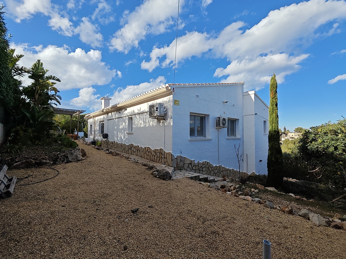 Villa exterior featuring white-washed walls with a natural stone base, set against a gravel path and lush Mediterranean gardens under a bright sky.