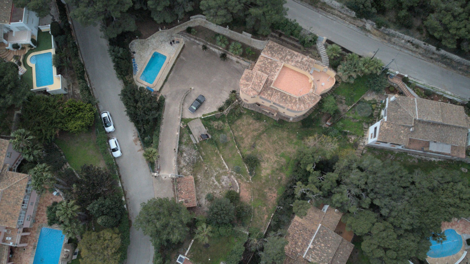 Aerial drone view showcasing the property layout, including the rectangular swimming pool, expansive paved driveway, and terraced Mediterranean garden.