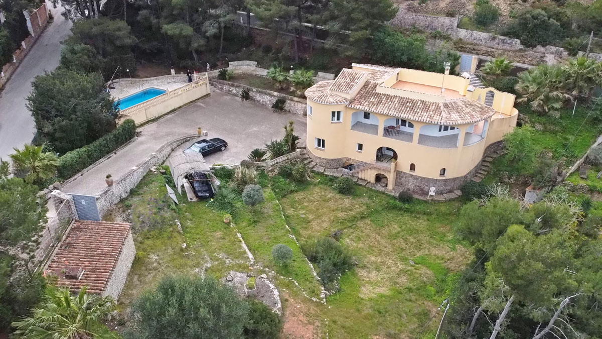 Aerial view of the Mediterranean villa featuring an arched terrace, stone-clad base, and tiered gardens. The property includes a private pool and a large paved driveway.