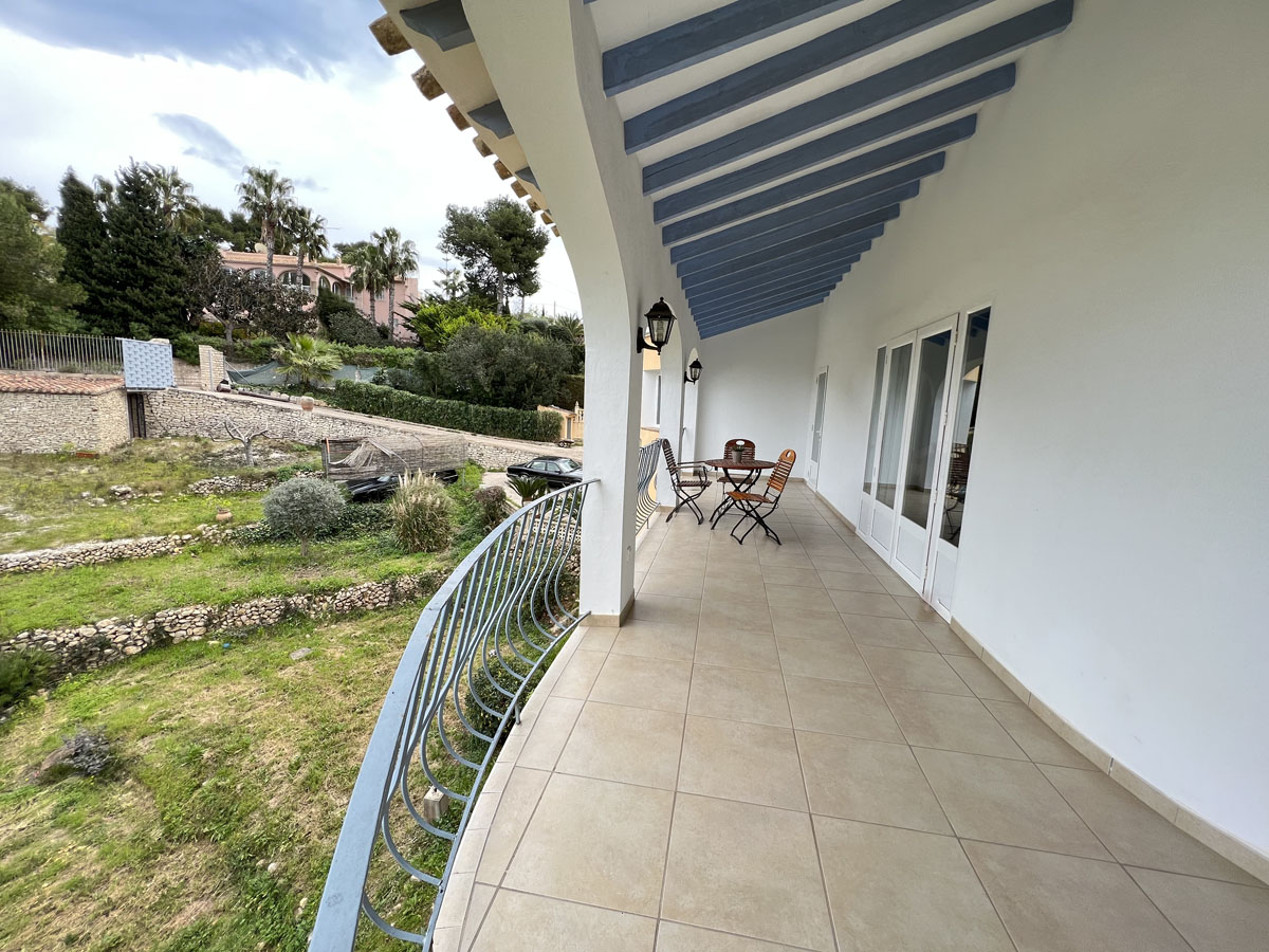 Covered terrace featuring blue wooden ceiling beams, tiled flooring, and a curved metal railing overlooking the terraced Mediterranean garden.