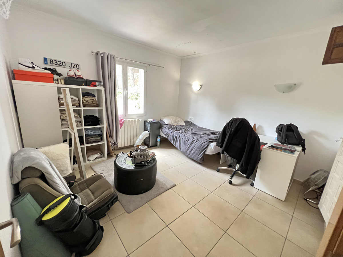 Independent studio bedroom featuring large ceramic floor tiles, white walls, and natural light. The layout offers space for a work desk and storage units.