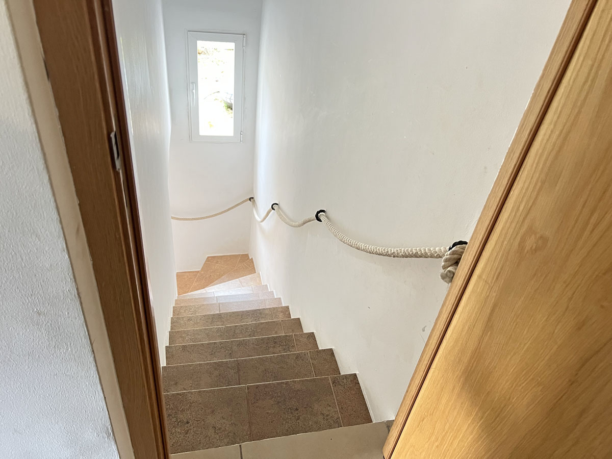 Internal staircase featuring ceramic tiled steps and a nautical rope handrail, illuminated by natural light from a slim vertical window.