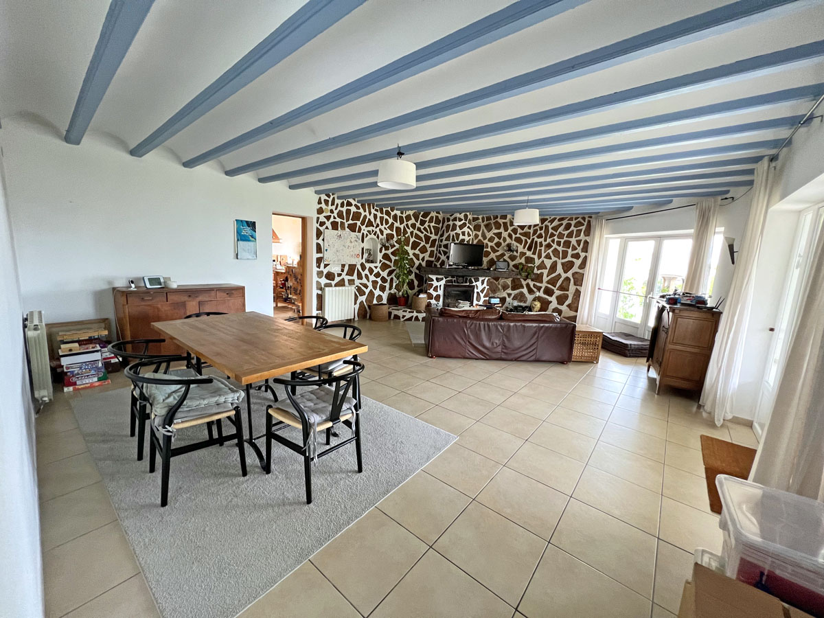 Open-plan living and dining area featuring blue-painted ceiling beams, a natural stone feature wall with a fireplace, and large ceramic floor tiles.