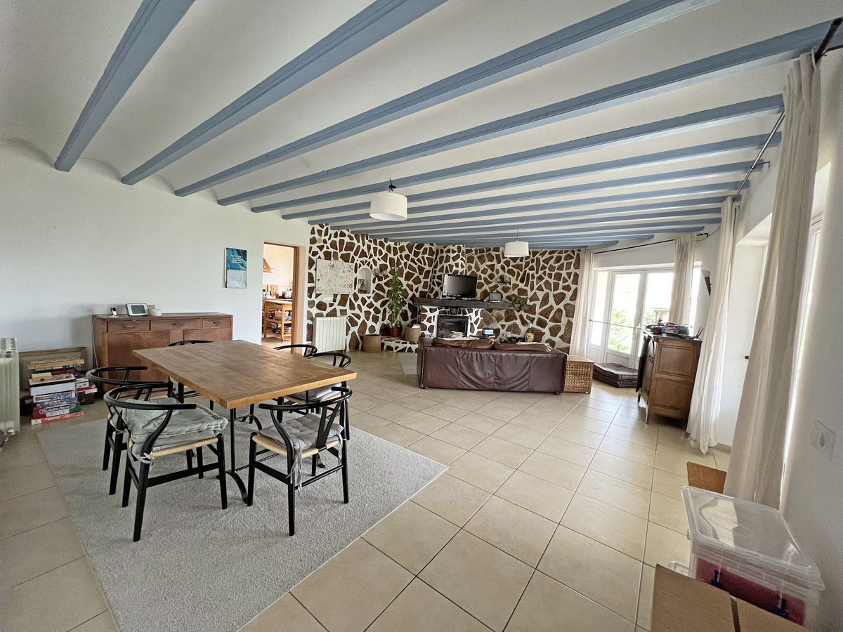Open-plan living and dining room featuring light blue exposed ceiling beams, a natural stone accent wall with a fireplace, and large ceramic floor tiles.