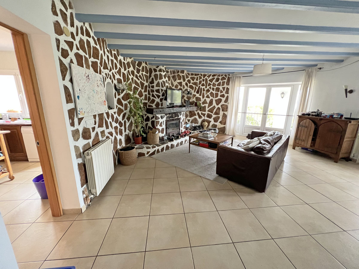 Rustic living room featuring a natural stone accent wall with integrated fireplace, light blue ceiling beams, and tiled flooring with terrace access.