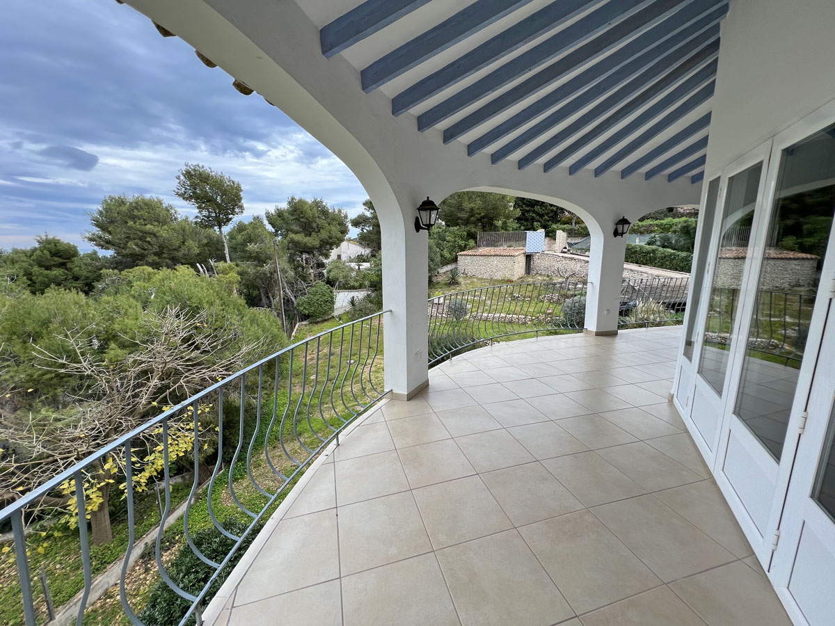 Spacious covered terrace with white arches, blue-painted ceiling beams, and tiled flooring overlooking the garden and Mediterranean Sea.