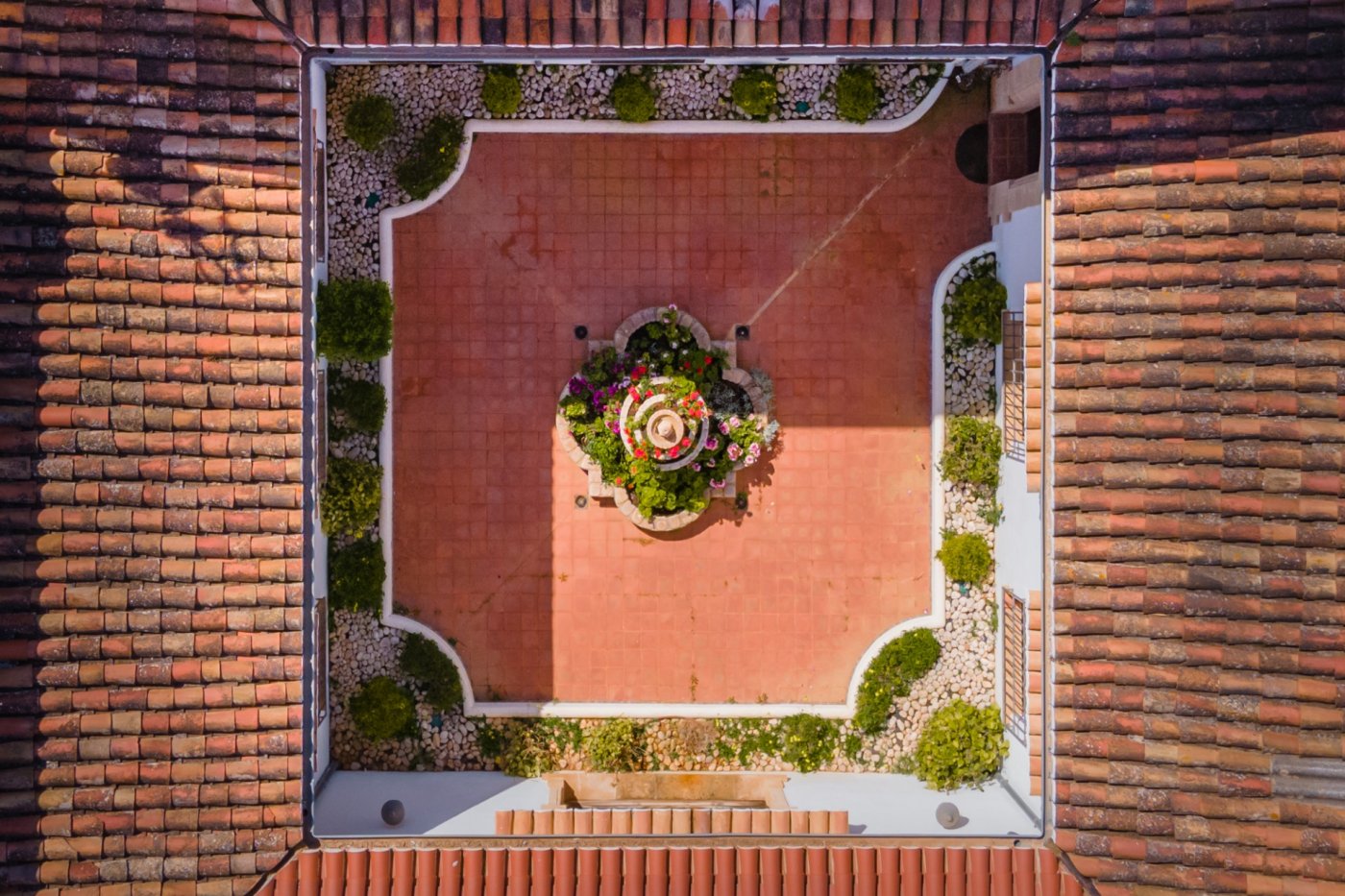 Aerial view of the central Mediterranean courtyard featuring terracotta tiling, a stone fountain with flowers, and perimeter landscaping framed by clay tile roofs.