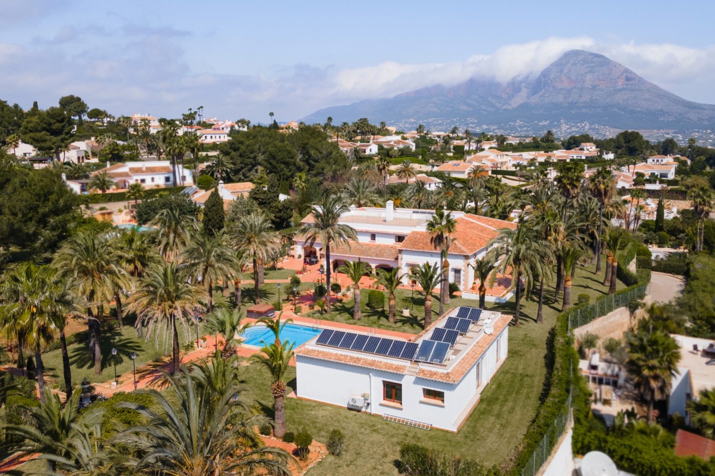 Aerial view of the estate featuring the main villa, guest house with solar panels, swimming pool, and extensive palm garden with Montgó mountain views.