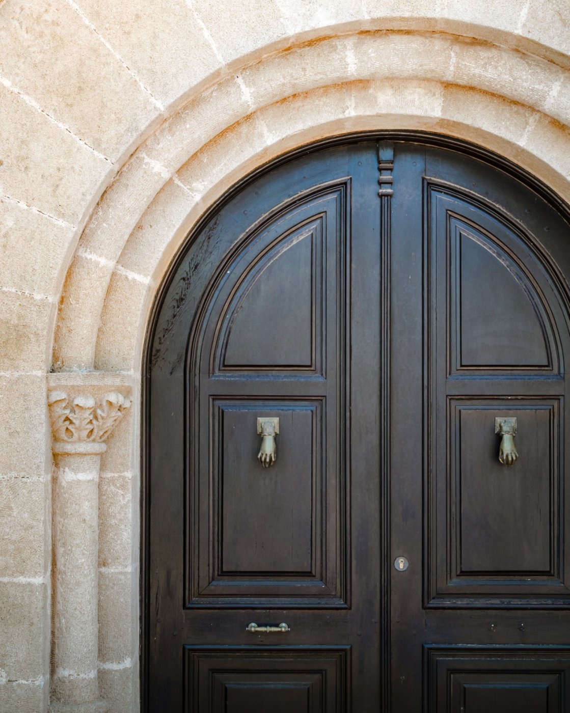 Arched dark wood entrance door set within a traditional Tosca stone archway, featuring ornate hand-shaped brass knockers and a carved column.