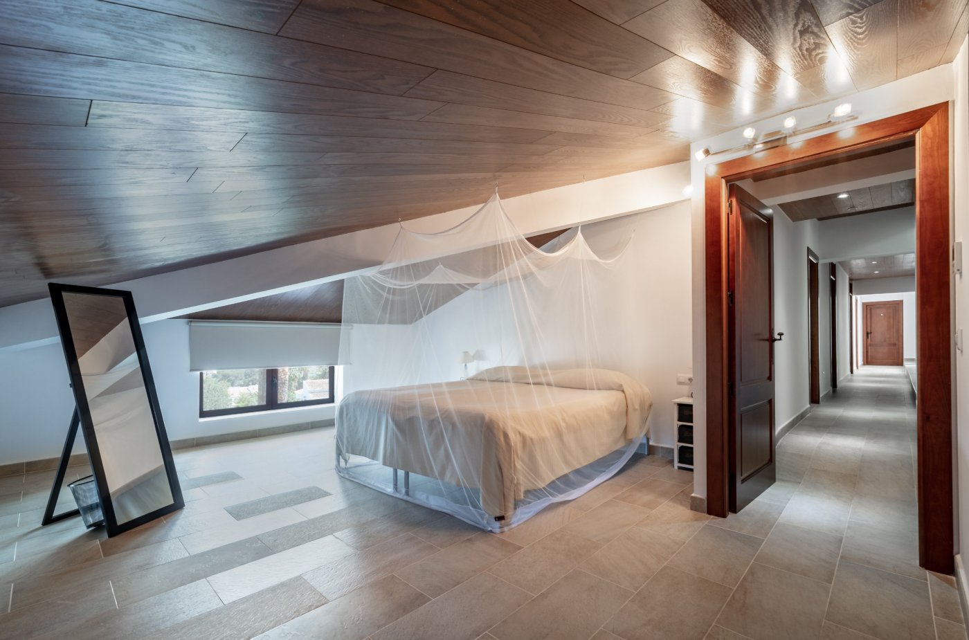 Attic bedroom featuring sloped wood-paneled ceilings, a bed with a mosquito net, and tiled flooring. The room includes a full-length mirror and opens to a corridor.