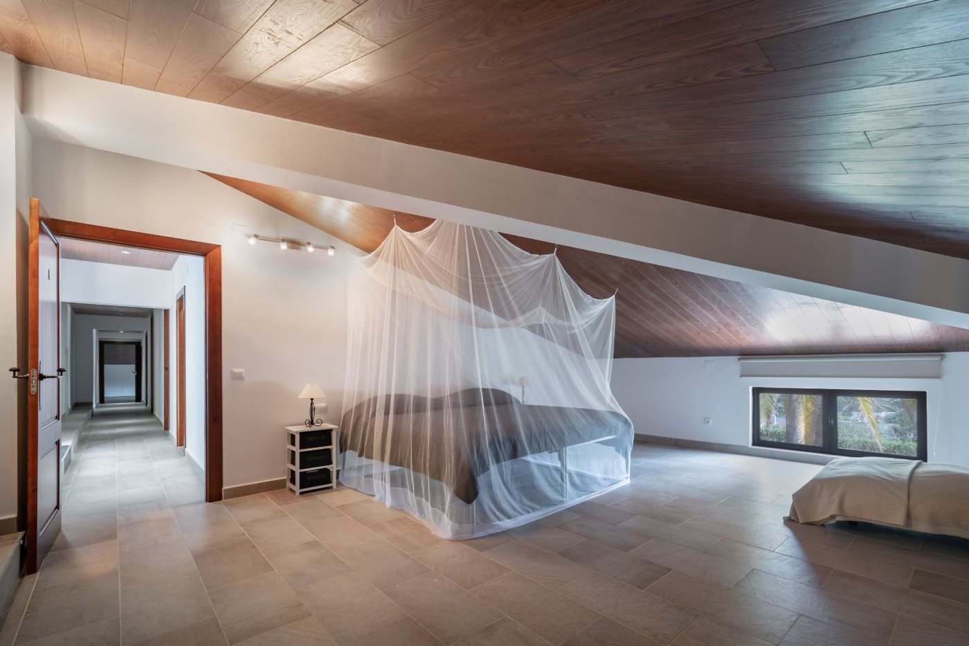 Attic bedroom featuring sloped wood-paneled ceilings, tiled flooring, and a bed with a mosquito net. The space includes a low window with views of the palm garden.