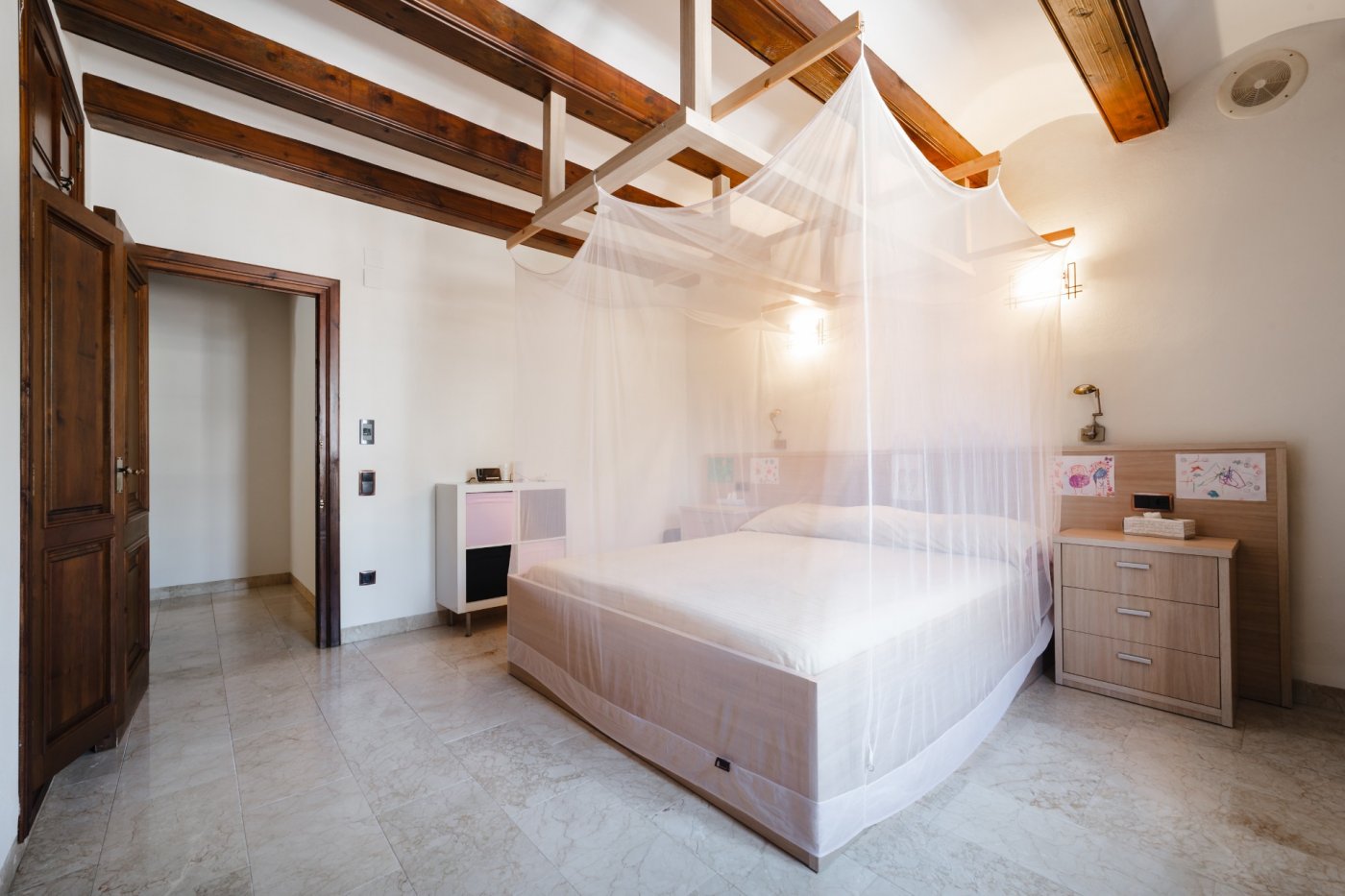 Bedroom featuring exposed dark wood ceiling beams, a bed with a mosquito net canopy, and polished stone flooring. Includes solid wood doors and wall lighting.