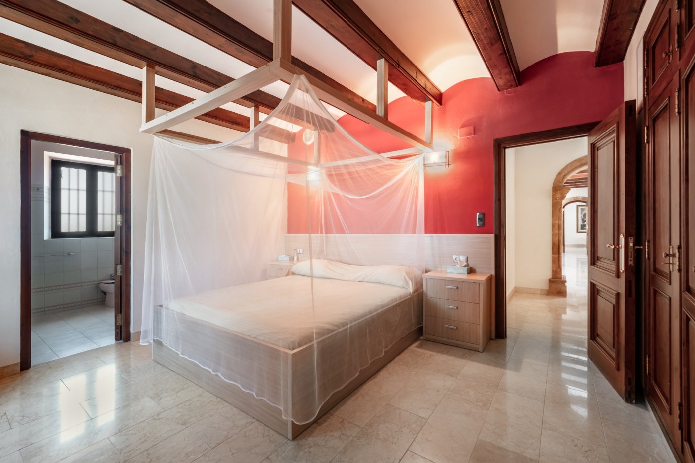 Bedroom featuring exposed wooden beams, a red accent wall, and a canopy bed. Includes marble flooring, an ensuite bathroom, and views of traditional stone arches.