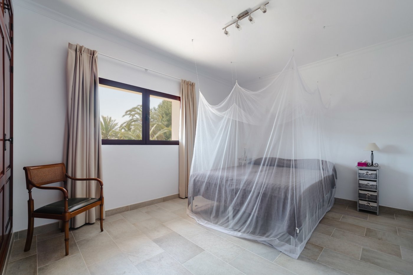 Bright bedroom featuring a double bed with a suspended mosquito net, tiled flooring, and a window overlooking the palm-filled garden. Includes classic wooden furniture.