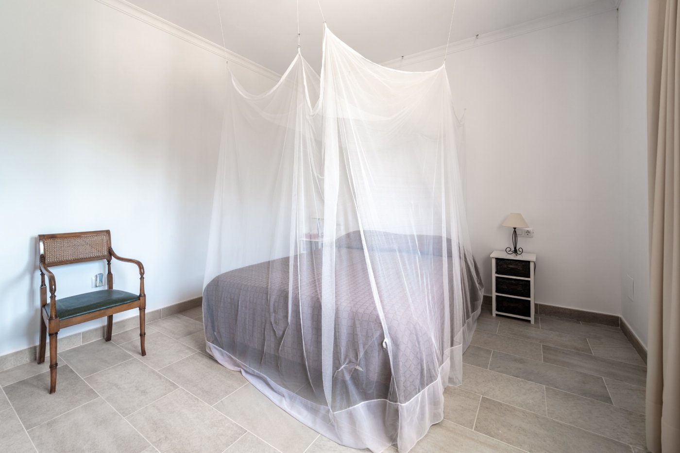Bright bedroom featuring stone-effect floor tiles, a double bed with a ceiling-mounted mosquito net, and elegant vintage-style furniture.