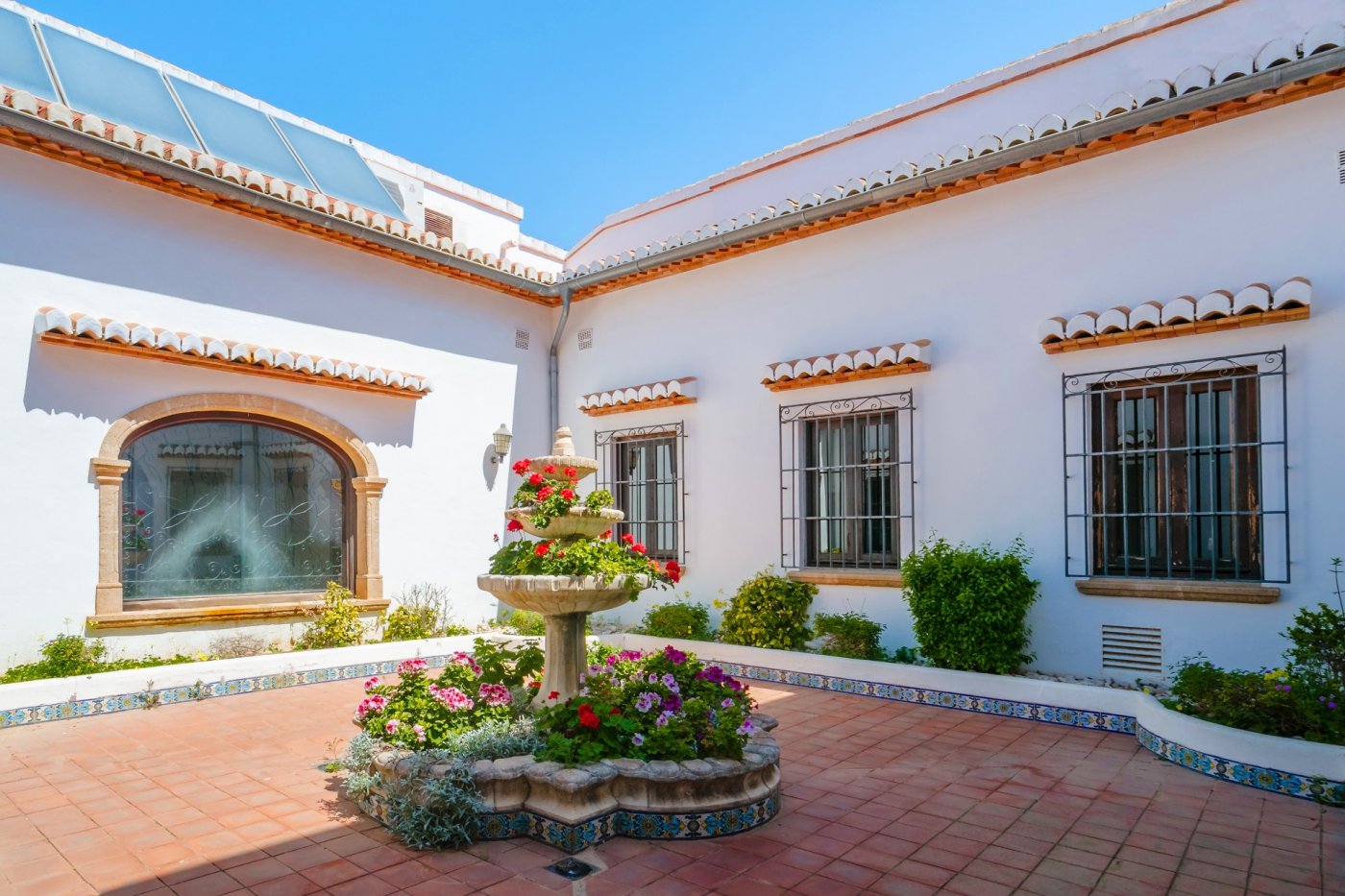 Central Mediterranean courtyard featuring a three-tiered stone fountain, terracotta tiling, and traditional Tosca stone arches under a clear sky.