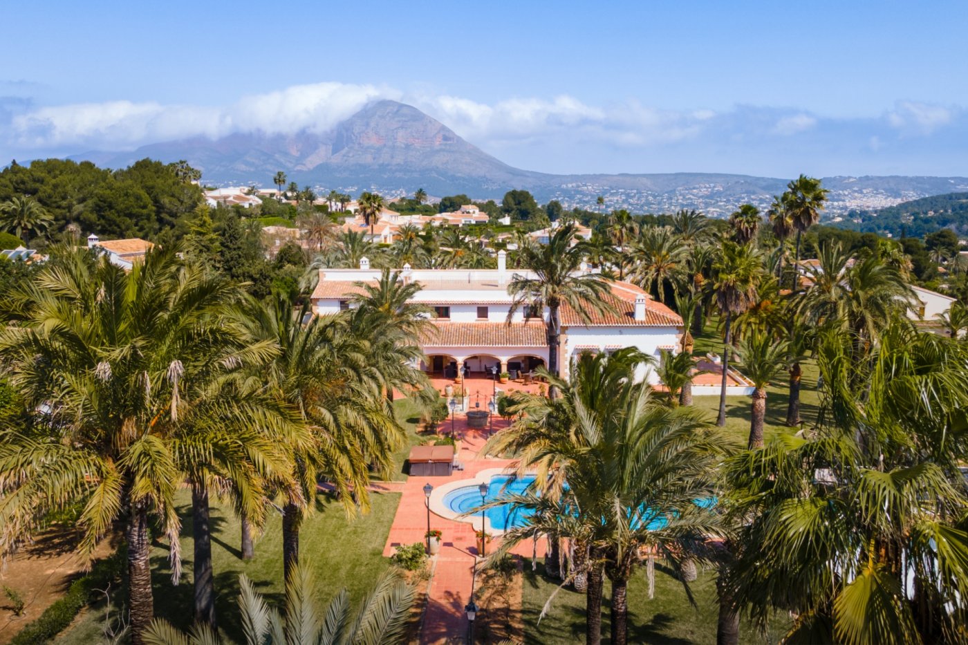 Elevated view of the Mediterranean villa featuring an expansive palm garden, a large swimming pool, and clear views of the Montgó mountain.