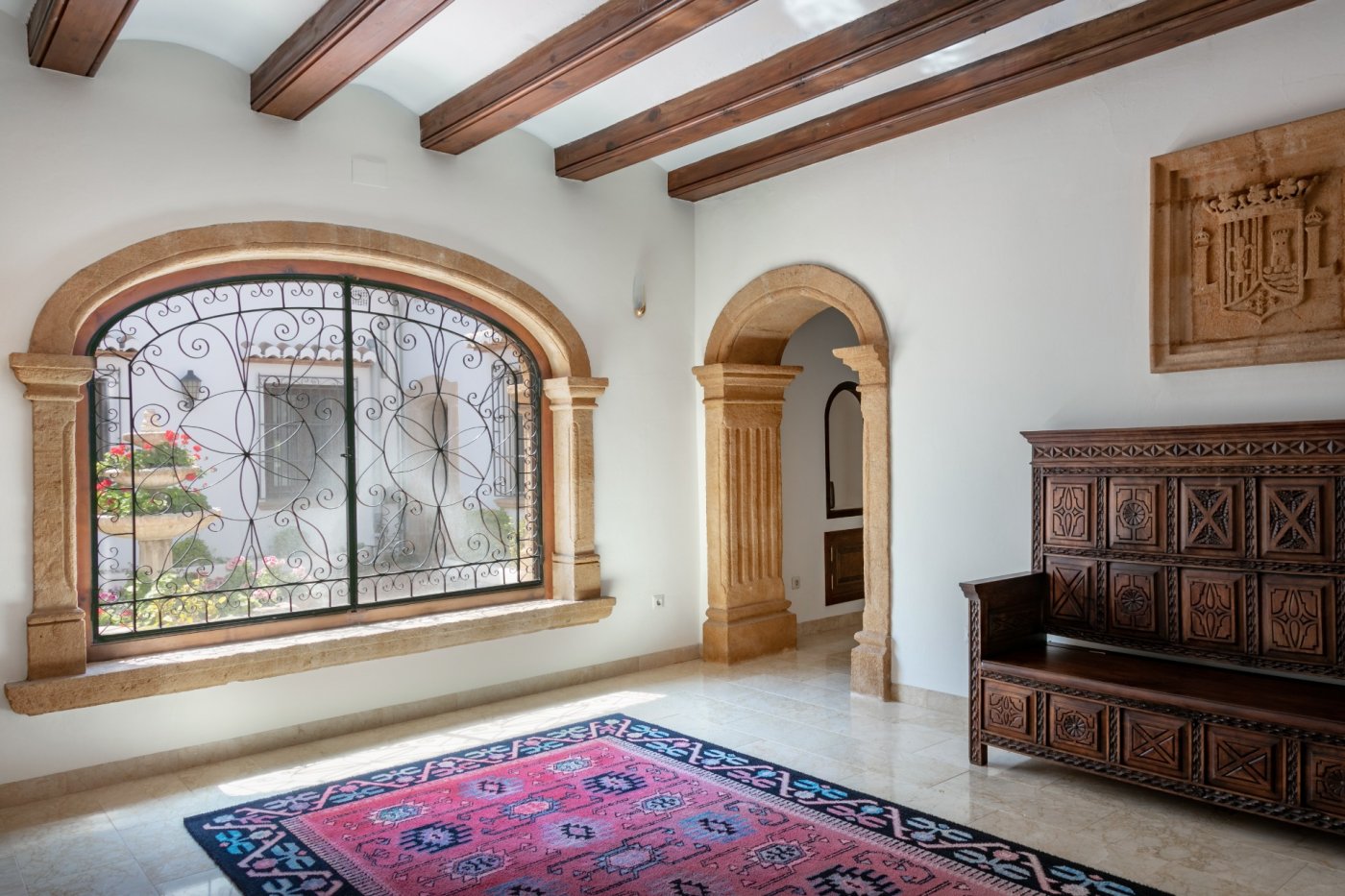 Entrance hall featuring traditional Tosca stone arches, exposed wooden beams, and a large window with wrought ironwork overlooking the courtyard fountain.
