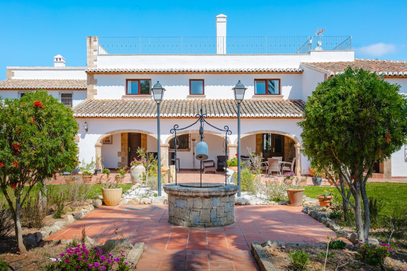 Exterior courtyard featuring a traditional stone well, terracotta tiling, and white facade with Tosca stone arches and Mediterranean shrubbery.