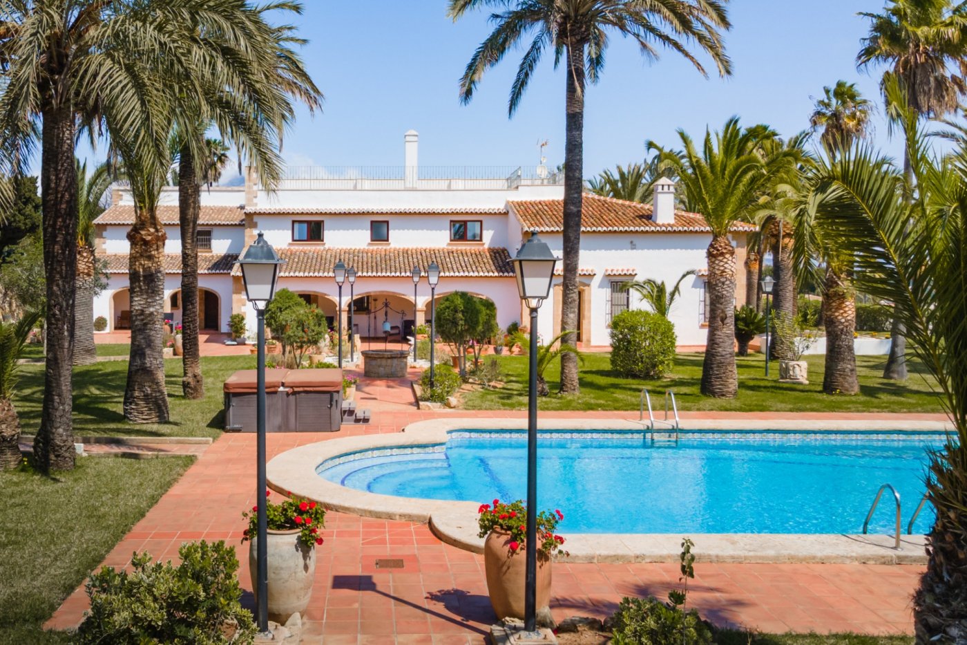 Exterior view of the Mediterranean villa featuring a large swimming pool, manicured lawn with mature palm trees, and a terracotta terrace with traditional lamp posts.