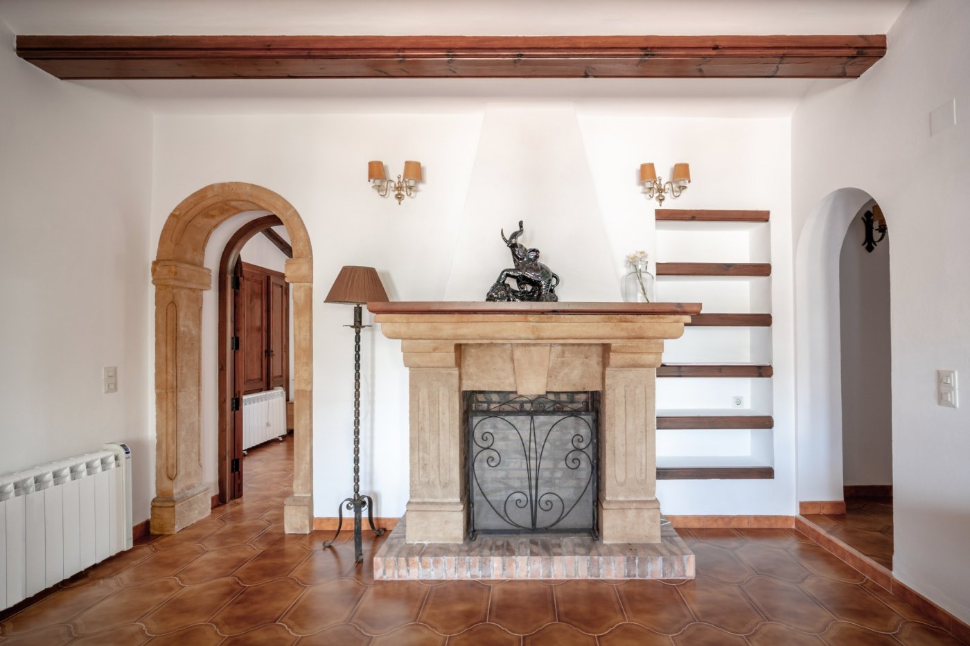 Living area featuring a hand-carved Tosca stone fireplace, exposed wooden beams, and a traditional archway. Terracotta flooring and built-in shelving enhance the rustic charm.