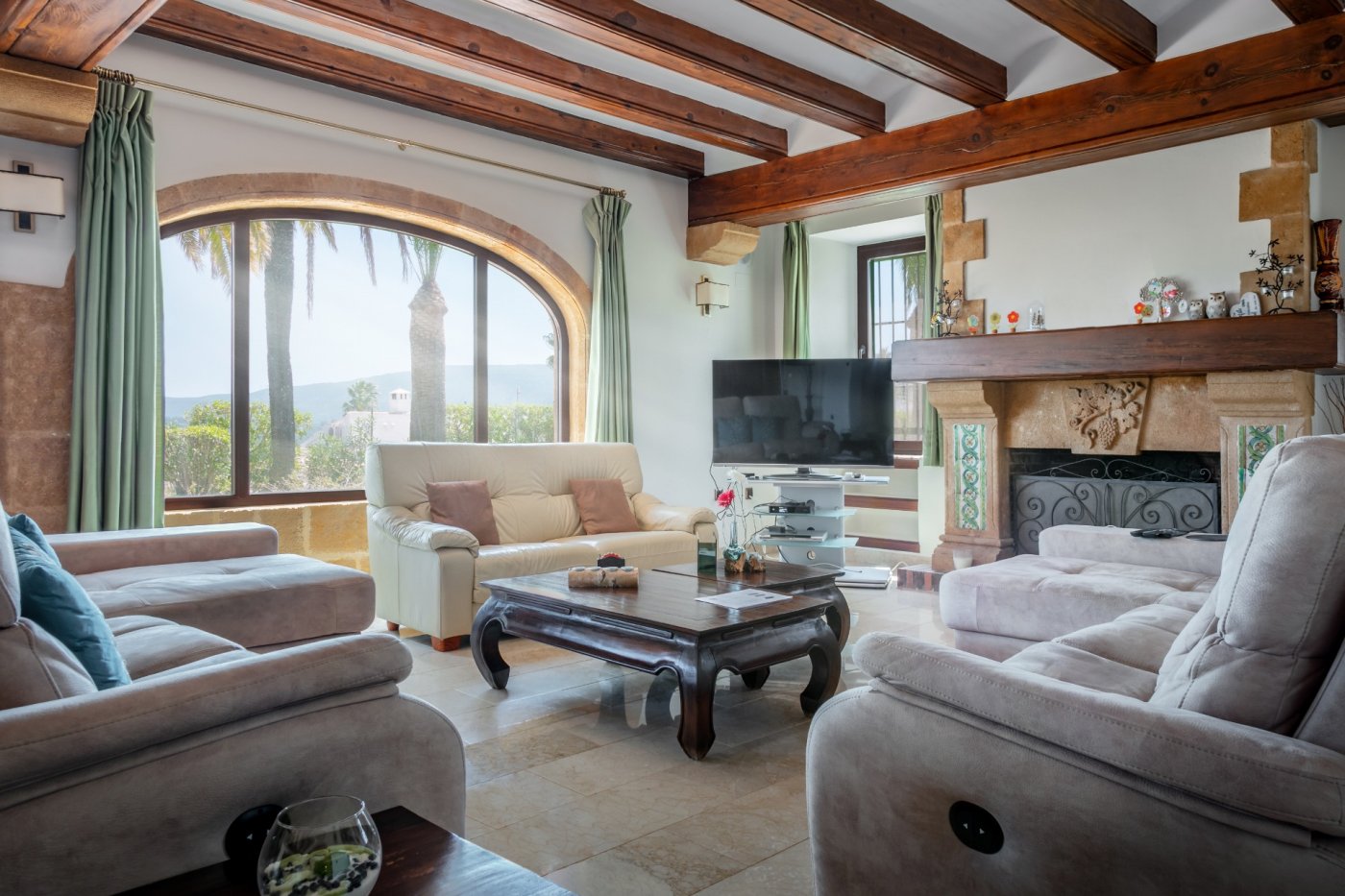 Living room featuring exposed wooden ceiling beams, a large Tosca stone arched window with garden views, and a traditional fireplace with decorative tiling.