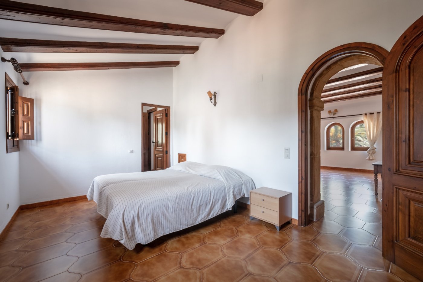 Mediterranean bedroom featuring exposed dark wood ceiling beams, rustic terracotta flooring, and a traditional Tosca stone archway leading to adjacent rooms.