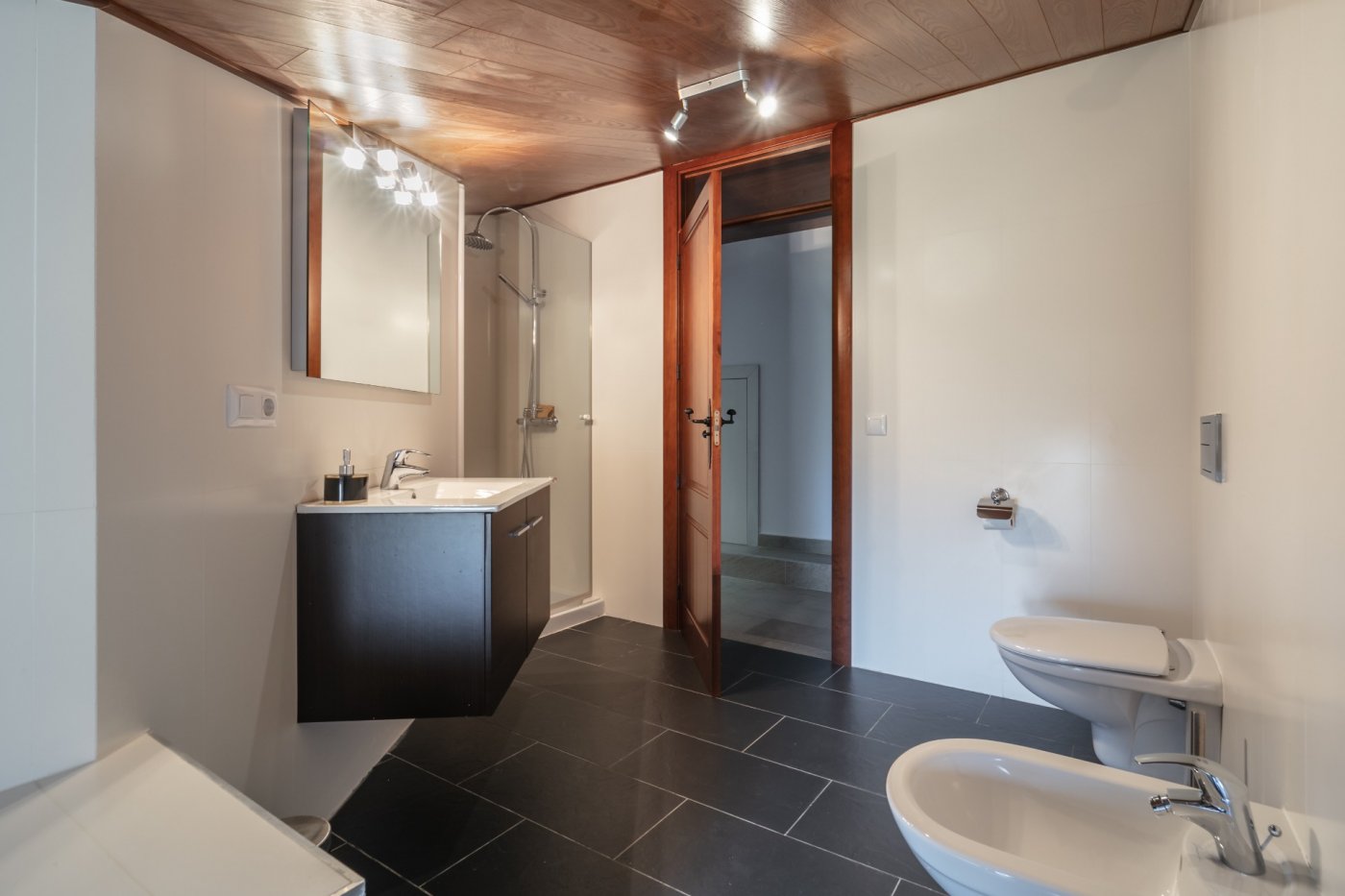 Modern bathroom featuring a wooden ceiling, dark slate flooring, a floating vanity, glass shower enclosure, and white porcelain fixtures.