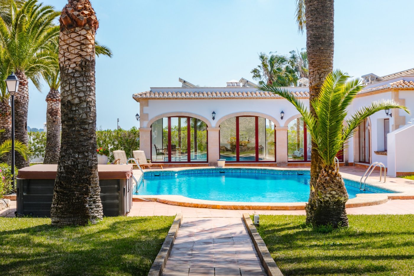 Outdoor swimming pool area with terracotta tiling, mature palm trees, a covered jacuzzi, and a villa featuring traditional stone arches and solar panels.