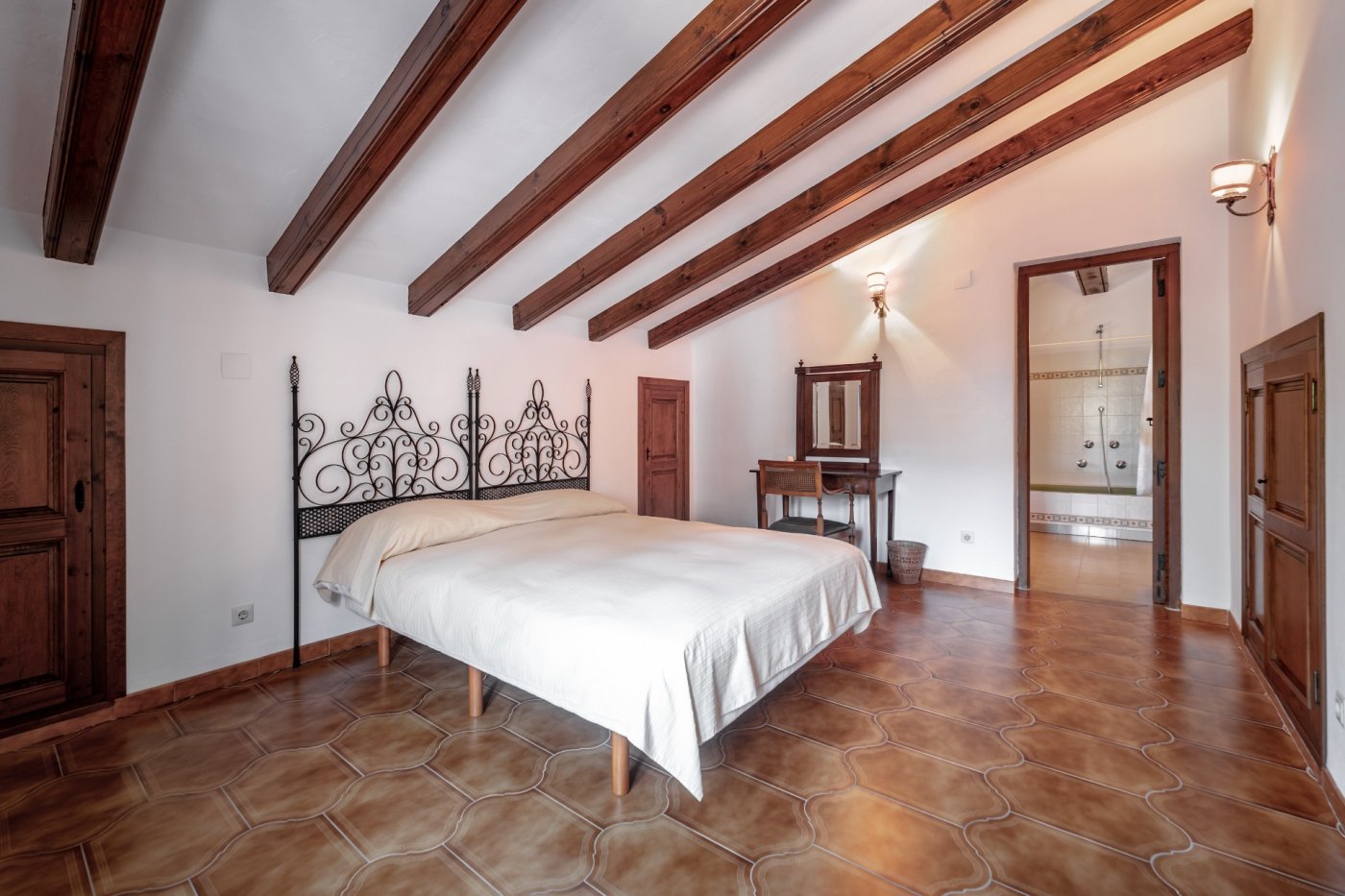 Rustic bedroom featuring exposed wooden ceiling beams, decorative terracotta flooring, a wrought iron headboard, and an integrated ensuite bathroom.