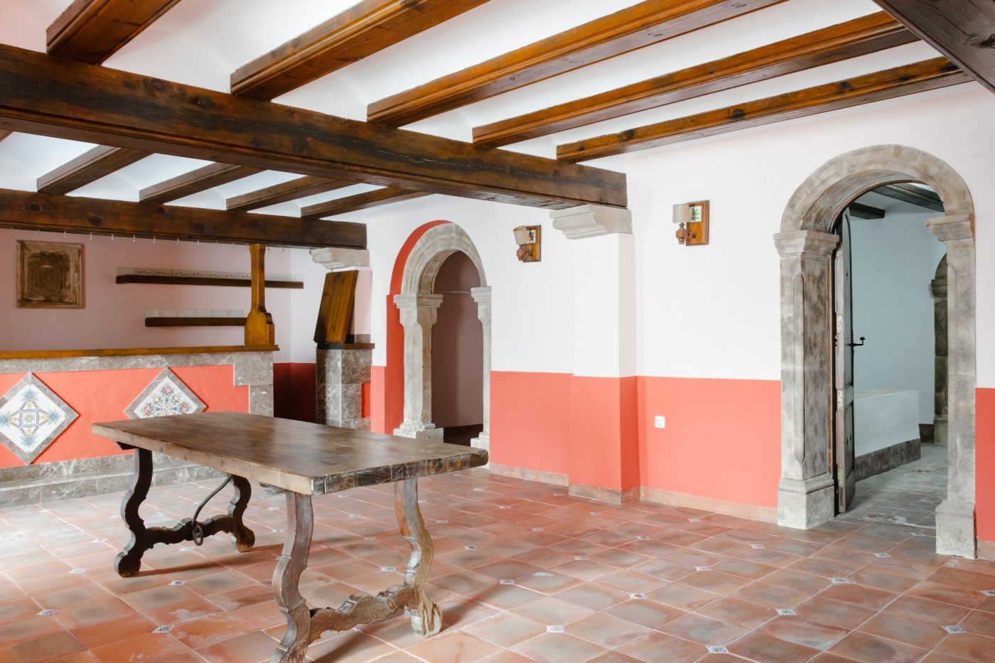 Rustic living area featuring exposed dark wood ceiling beams, terracotta tile flooring, and hand-carved Tosca stone arches. A solid wood table complements the traditional decor.